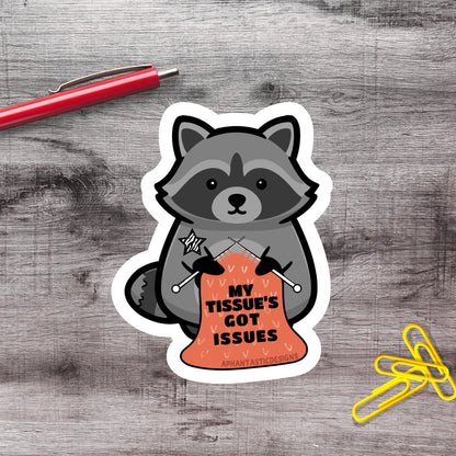 A sticker depicting a raccoon holding a sign that says "My Tissue's Got Issues", with a red pen and yellow paper clips on a wooden surface.