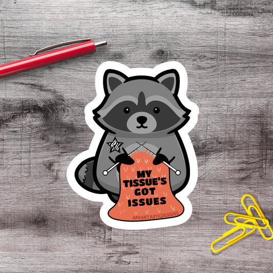 A sticker depicting a raccoon holding a sign that says "My Tissue's Got Issues", with a red pen and yellow paper clips on a wooden surface.