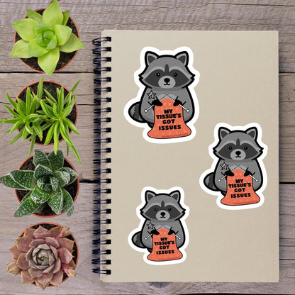 a spiral notebook with stickers of raccoons holding signs that read "My Tissue's Got Issues", accompanied by three potted succulents on a wooden surface.
