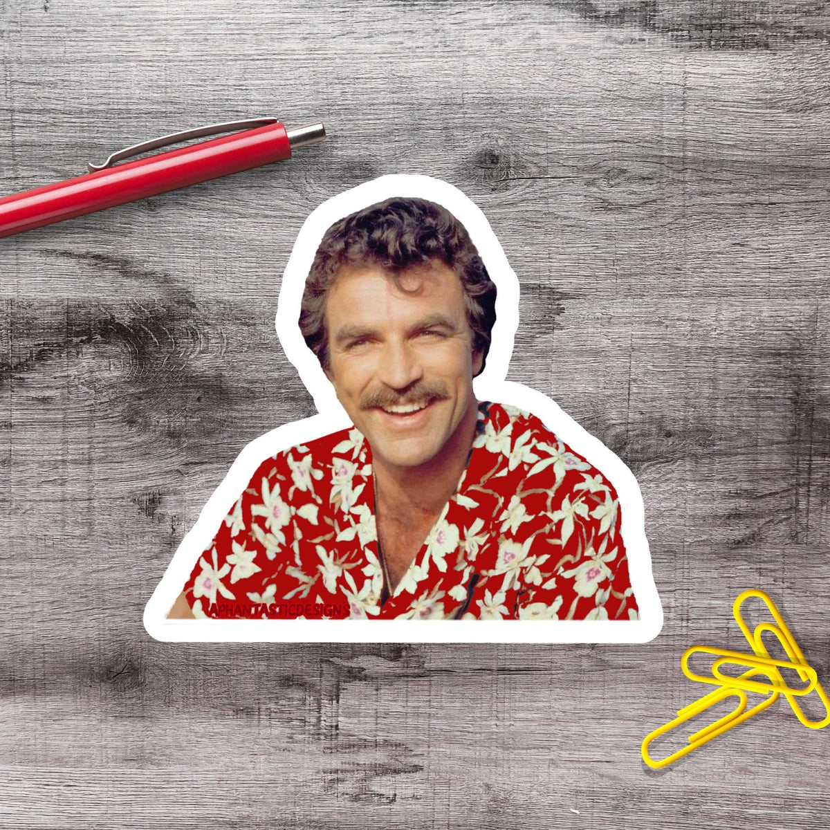 A sticker of a smiling man wearing a red and white floral shirt, placed on a wooden surface next to a red pen and yellow paper clips.