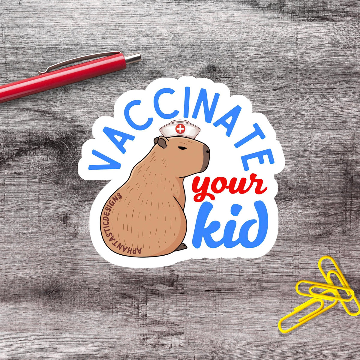A sticker with a cartoon image of a capybara wearing a nurse's hat and the text "VACCINE YOUR KID" on a wooden surface.